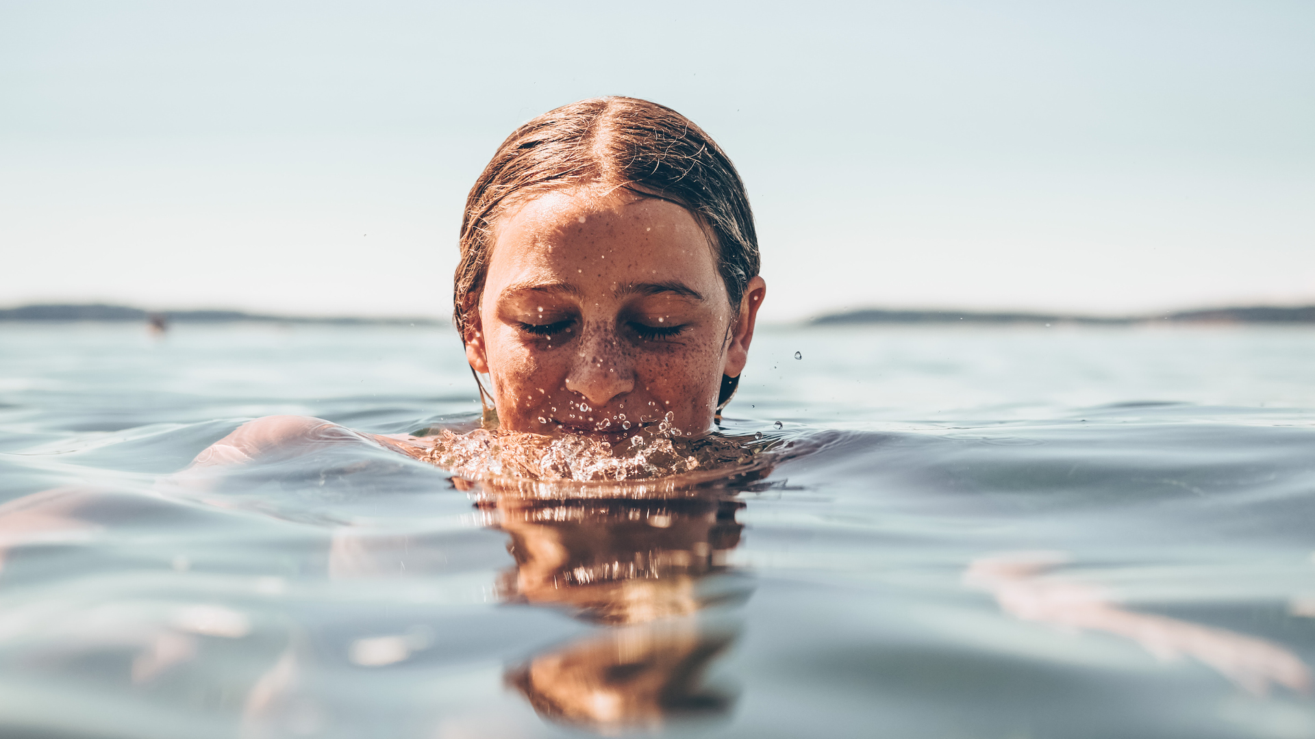 Der Kopf einer junge Frau mit Sommersprossen taucht aus dem Wasser auf. Sie bläst Luftblasen ins Wasser.