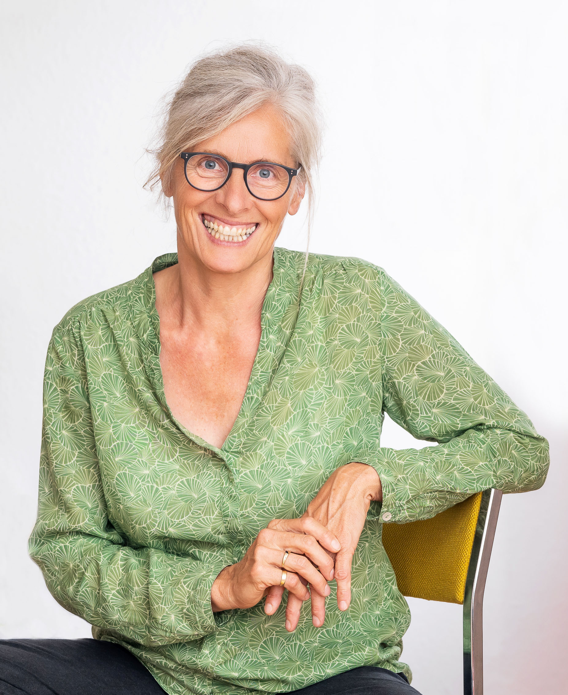 Gitte Sohler sitzt lächelnd auf einem Stuhl mit grüner Bluse und Brille.